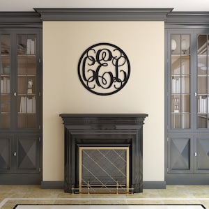 May include: A circular black wall hanging featuring an intricate, interconnected monogram design with three stylised letters. The piece is constructed from a thin, flat material and is mounted centrally on a light-coloured wall above a fireplace.