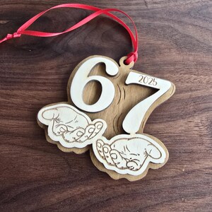 May include: Wooden ornament with the numbers 6 and 7, and the year 2025. The ornament has a red ribbon for hanging and features two hands cradling a baby. The ornament is made of light brown wood.