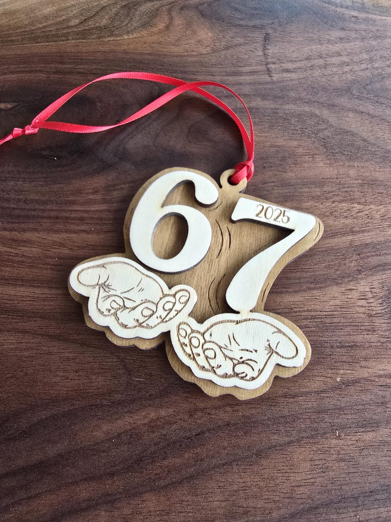 May include: Wooden ornament with the numbers 6 and 7, and the year 2025. The ornament has a red ribbon for hanging and features two hands cradling a baby. The ornament is made of light brown wood.