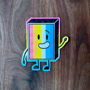 May include: A colorful sticker of a cartoon character with a smiling face. The character is a rectangle with a yellow, white, and blue striped pattern. The character has black eyes, a black outline, and a black arm raised in a wave. The sticker has a glittery finish.