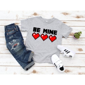 May include: A gray t-shirt with the text "BE MINE" in black letters and three red pixelated hearts.