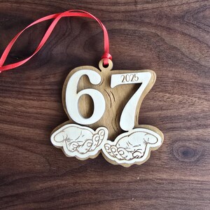 May include: Wooden ornament with the numbers "67" and the year "2025" cut out. The ornament has a red ribbon for hanging and features two hands. The ornament is made of light brown wood.