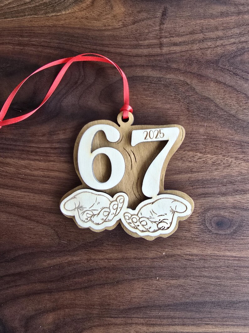 May include: Wooden ornament with the numbers "67" and the year "2025" cut out. The ornament has a red ribbon for hanging and features two hands. The ornament is made of light brown wood.