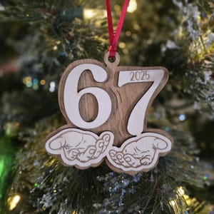 May include: Wooden Christmas ornament with the numbers "67" and "2025" cut out. The ornament features a wood grain background and a red ribbon for hanging. The bottom of the ornament has a graphic of two hands.