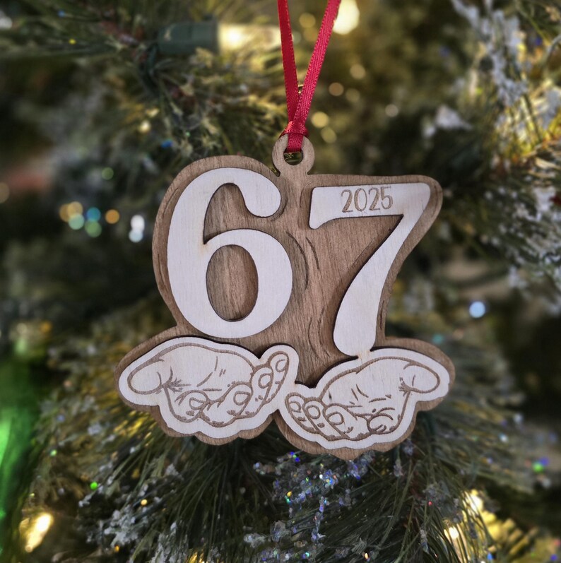 May include: Wooden Christmas ornament with the numbers "67" and "2025" cut out. The ornament features a wood grain background and a red ribbon for hanging. The bottom of the ornament has a graphic of two hands.