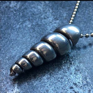 May include: A silver spiral shell pendant necklace. The shell is made of smooth, rounded, silver beads that are connected to form a spiral shape. The necklace is on a silver chain.