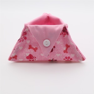 May include: A pink fabric pouch with a white button closure. The pouch is decorated with a pattern of paw prints, hearts, and bones.