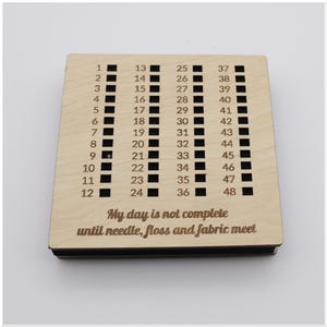 May include: A wooden needle minder with a grid of 48 numbered slots. The text "My day is not complete until needle, floss and fabric meet" is engraved on the surface. The minder is light brown.