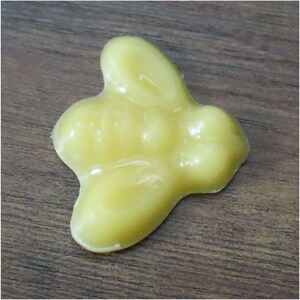 May include: A light yellow, bunny-shaped wax melt. The wax melt has a smooth, glossy surface and is sitting on a dark brown wooden surface. The bunny shape has rounded features.