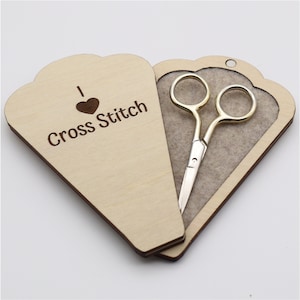 May include: A wooden box with a heart and the text "I Love Cross Stitch" engraved on the front. The box is open and a pair of small gold scissors are inside.