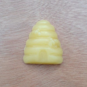 May include: A light yellow, beehive-shaped candle. The candle has a textured surface with horizontal ridges and a small arched doorway at the base. The candle is set against a light brown wooden background.