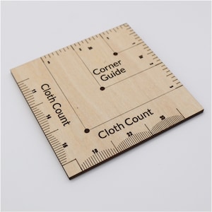 May include: A wooden square ruler with markings for measuring fabric. It has two sets of numbers, one for "Cloth Count" and one for "Corner Guide".