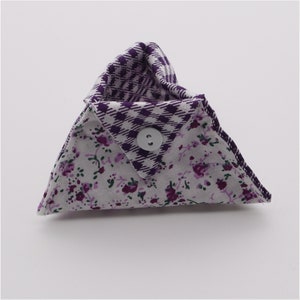 May include: A small, triangular fabric pouch with a floral pattern in white and purple, and a purple and white checkered interior. The pouch is secured with a gray button closure. The edges are neatly sewn, and the overall design is compact and decorative.