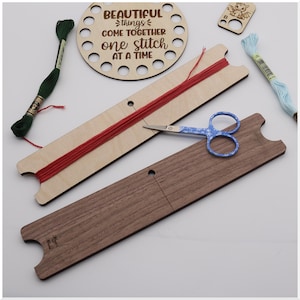 May include: A wooden embroidery floss organizer with red thread, a pair of blue patterned scissors, and a wooden circular piece with the text "BEAUTIFUL things COME TOGETHER one stitch AT A TIME". Also shown are spools of green and light blue embroidery floss.