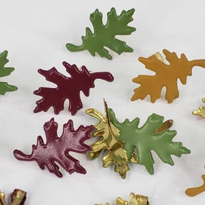 May include: A collection of leaf-shaped pins in various colors, including green, burgundy, and gold. The pins are arranged on a white surface, showcasing their autumnal theme. These decorative pins are ideal for adding a touch of nature to clothing or accessories.