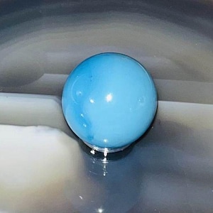 May include: A smooth, light blue sphere made of glass or ceramic. The sphere is sitting on a gray and white surface.