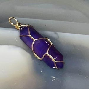 Puede incluir: Un colgante de cristal hexagonal alargado de color morado intenso, envuelto en un fino alambre de color dorado. El cristal tiene una punta afilada y una base plana, con el alambre formando una jaula decorativa alrededor de la piedra.