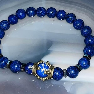 May include: A beaded bracelet featuring round, deep blue speckled beads and small, dark metallic cube spacers. A central, larger blue and white speckled bead is encased in an ornate, antique-style brass crown-shaped metal setting.
