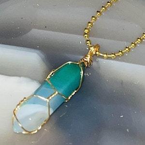 Könnte beinhalten: Goldfarbene Halskette mit Anhänger. Der Anhänger ist ein Kristall in Türkis-, Blau- und Weißtönen, umwickelt mit goldenem Draht. Die Halskette wird vor einem grau-weißen Hintergrund präsentiert.