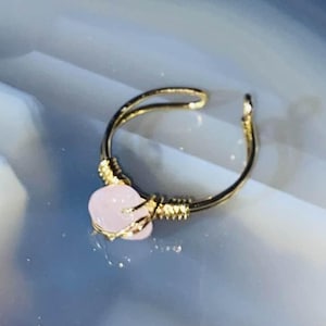 May include: A gold-colored adjustable ring with a light pink, heart-shaped stone. The ring's band has a twisted design and the stone is secured with gold-colored prongs. The ring is set against a light blue and white background.