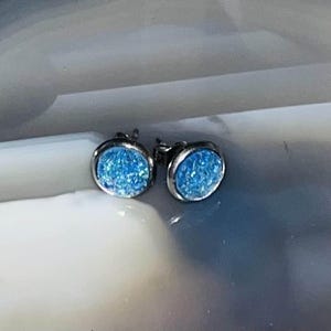 Może przedstawiać: Para srebrnych kolczyków z jasnoniebieskimi akcentami brokatowymi.