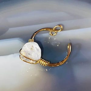 Könnte beinhalten: Ein goldfarbener Ring mit einem klaren, unregelmäßig geformten Stein. Der Stein wird von einem filigranen Drahtkäfig gehalten. Das Band des Rings verläuft um den Stein herum, mit einem Schlaufendesign an der Oberseite.