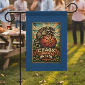 May include: A navy blue garden flag features a raccoon perched on a basketball. The flag's design includes the words "Chaos Bracket Energy" in a vintage style. The flag is displayed on a black metal stand in a backyard setting.