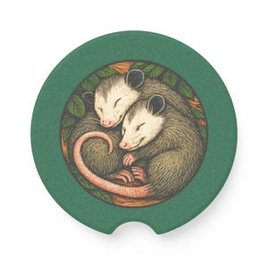 May include: A green car coaster featuring a detailed illustration of two opossums curled up together. The opossums are gray and white with pink noses and ears, surrounded by green leaves and a brown border.