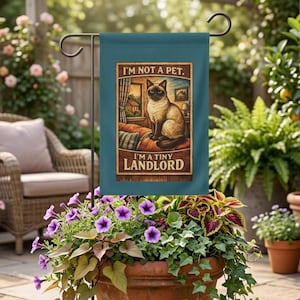 Può includere: Una bandiera da giardino color verde acqua con un gatto siamese seduto su un divano e il testo I'M NOT A PET. I'M A TINY LANDLORD. La bandiera è su un supporto in metallo nero e mostra un'illustrazione dettagliata di un gatto in una stanza.