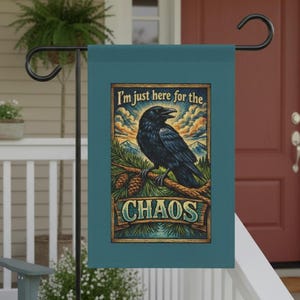 Puede incluir: Bandera de jardín de color verde azulado con una ilustración detallada de un cuervo negro posado en una rama con piñas. La bandera tiene el texto "I'm just here for the CHAOS" en estilo vintage.