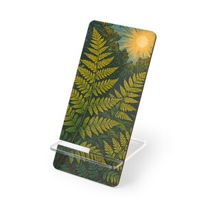 Puede incluir: Un soporte para teléfono con un diseño de helecho, un sol amarillo y follaje verde. El soporte es de acrílico transparente y sostiene un teléfono o tableta rectangular. La obra de arte tiene un estilo de ilustración botánica vintage.