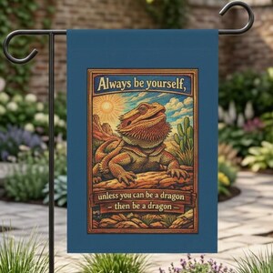 Puede incluir: Una bandera de jardín azul con una ilustración de un dragón barbudo en un paisaje desértico. La bandera tiene el texto "Always be yourself, unless you can be a dragon - then be a dragon". Está colgada de un soporte de metal negro.