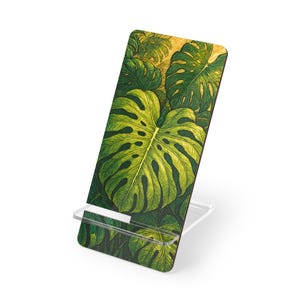 May include: A wooden phone stand featuring a vibrant illustration of large, green monstera leaves. The leaves have intricate details and varying shades of green. The stand has a clear, acrylic base, providing support for a phone or tablet.