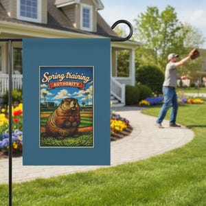 Könnte beinhalten: Eine blaue Gartenflagge mit der Aufschrift "Spring Training Authority" und einer Illustration eines Murmeltiers auf einem Baseballfeld. Die Flagge wird auf einem schwarzen Metallständer in einem Garten mit einem Haus im Hintergrund präsentiert.