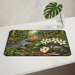 Puede incluir: Una alfombrilla de escritorio rectangular con una colorida ilustración de una zarigüeya durmiendo bajo flores blancas en un bosque. Un pequeño arroyo fluye a través del bosque, con dos ciervos de pie al fondo entre árboles y follaje.