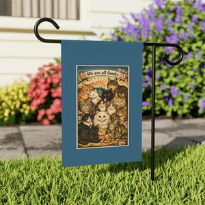 May include: A blue garden flag with a cat-themed illustration and the words "We are all family." The design includes a mountain and sun. The flag is displayed on a black metal stand in a garden.