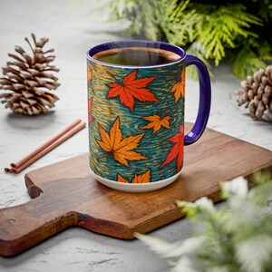 Puede incluir: Una taza de cerámica con un vibrante diseño de hojas de otoño. La taza presenta hojas de arce naranjas y rojas sobre un fondo turquesa, con un asa y borde morados. La taza está llena de un líquido oscuro, posiblemente café o té.