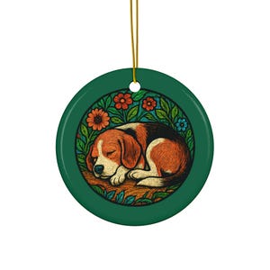 May include: A round, green ceramic ornament featuring a sleeping beagle dog illustration. The dog is brown, black, and white, surrounded by orange and blue flowers and green leaves. A gold string is attached for hanging.