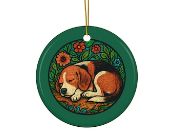 Op de afbeelding: Een ronde, groene keramische ornament met een illustratie van een slapende beagle hond. De hond is bruin, zwart en wit, omringd door oranje en blauwe bloemen en groene bladeren. Een gouden touwtje is bevestigd om op te hangen.