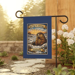 May include: A navy blue garden flag featuring a groundhog in a snowy landscape with the text "Spring is coming... eventually." The illustration has a vintage style with a sunrise in the background. The flag is displayed on a black metal stand.