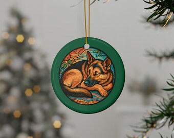 May include: A round, green ceramic ornament featuring a stylized illustration of a German Shepherd dog. The dog is depicted in shades of brown, black, and orange, resting within a colorful, circular design. A gold string is attached for hanging.