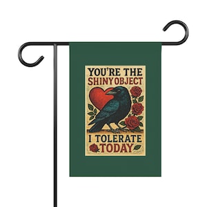 May include: A dark green garden flag with a black metal stand. The flag features a raven illustration with a red heart and roses, and the text "YOU'RE THE SHINY OBJECT I TOLERATE TODAY."