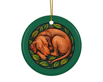 Op de afbeelding: Een ronde, groene keramische ornament met een illustratie van een slapende bruine hond omringd door groene bladeren. Het ornament heeft een gouden touwtje om op te hangen. De hond is opgerold, met gesloten ogen.