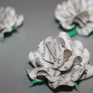 May include: Close-up of handmade paper flowers with green paper leaves. The flowers are made from pages of a book, with visible text. The flowers are arranged on a gray surface.