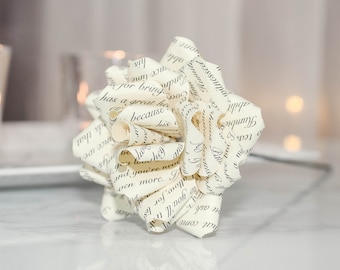 May include: A decorative paper flower made from pages of a book, with visible text. The flower is cream-colored with black text and is in focus. Two glass candle holders with lit candles are in the background.