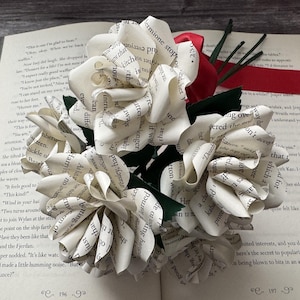 May include: A bouquet of paper flowers made from book pages, with green stems and a red ribbon. The flowers are white with visible text, resting on an open book. A unique, literary-themed decorative item.