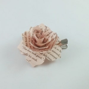 May include: A handmade paper rose hair clip. The rose is crafted from pages of a book, with visible text adding a unique touch. The petals are a soft pink hue, and the clip is silver-toned. A delicate and literary accessory.