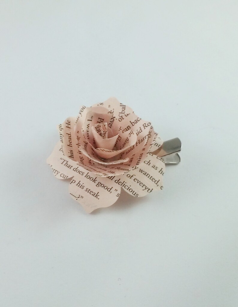 May include: A handmade paper rose hair clip. The rose is crafted from pages of a book, with visible text adding a unique touch. The petals are a soft pink hue, and the clip is silver-toned. A delicate and literary accessory.