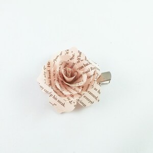 May include: A handmade paper rose hair clip. The rose is crafted from pages of a book, with visible text. The petals are a soft pink hue, and the clip is silver-toned. A unique accessory for hair styling.