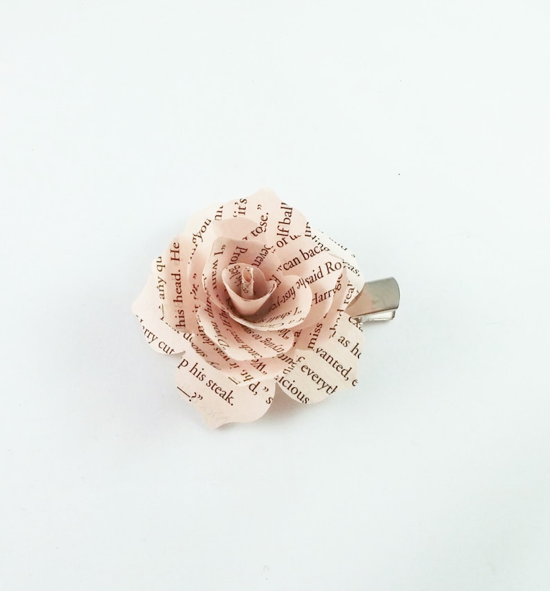 May include: A handmade paper rose hair clip. The rose is crafted from pages of a book, with visible text. The petals are a soft pink hue, and the clip is silver-toned. A unique accessory for hair styling.